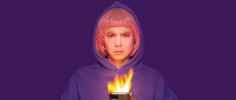 Die Schauspielerin Anastasia Lara Heller steht vor einem violetten Hintergrund. Sie trägt eine pinke Perücke, einen lilafarbenen Hoodie und hat die Kapuze auf. Sie hält ein Handy in beiden Händen, das brennt.