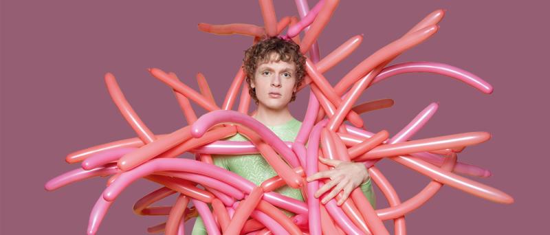 Ein junger Mann steht vor einem altrosafarbenen Hintergrund. Er hat einen ernsten Gesichtsausdruck. Er trägt ein hellgrünes T-Shirt und ein Kostüm, dass aus lauter rosafarbenen und orangen Modellier-Luftballons besteht.