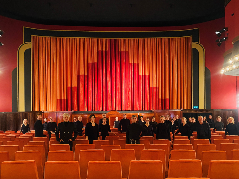 Das Bild zeigt den Chor der Hochschule für bildende Künste. Die Personen stehen im Zuschauerraum eines Theaters, hinter ihnen die Bühne mit rotem Vorhang.