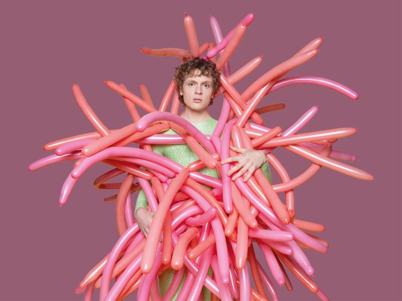 Ein junger Mann steht vor einem altrosafarbenen Hintergrund. Er hat einen ernsten Gesichtsausdruck. Er trägt ein hellgrünes T-Shirt und ein Kostüm, dass aus lauter rosafarbenen und orangen Modellier-Luftballons besteht.