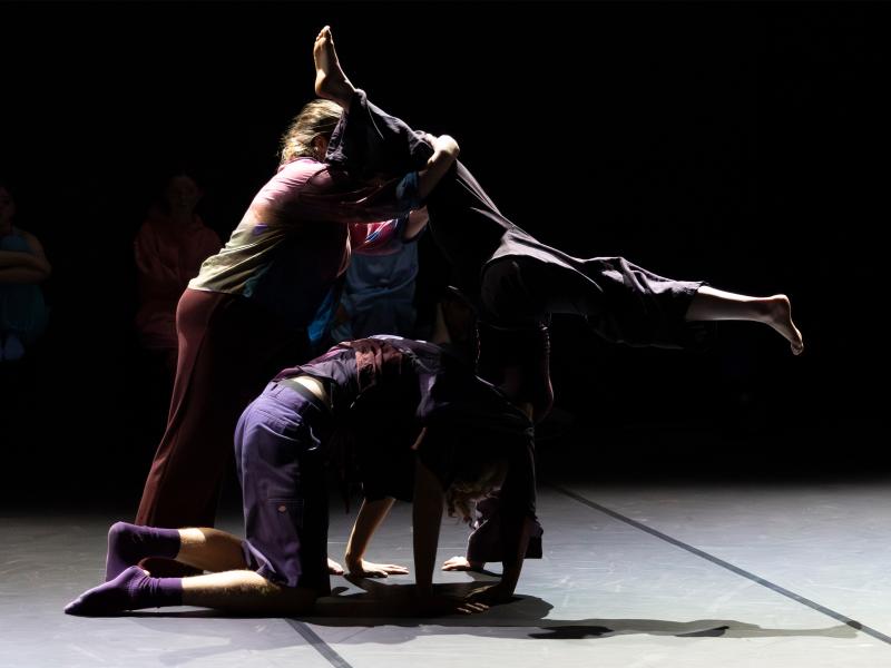 Eine Gruppe von Tänzer*innen, die eng aneinander verschiedene Bewegungen machen. Eine Tänzerin macht einen Spagat im Handstand, während eine andere eines ihrer Beine festhält. 