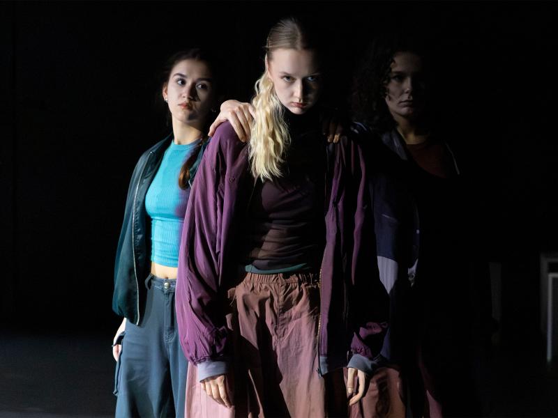 Zwei Tänzerinnen stehen hinter einer weiteren und haben ihre Hände auf ihren Schultern liegen. Sie schauen alle nach vorne. 