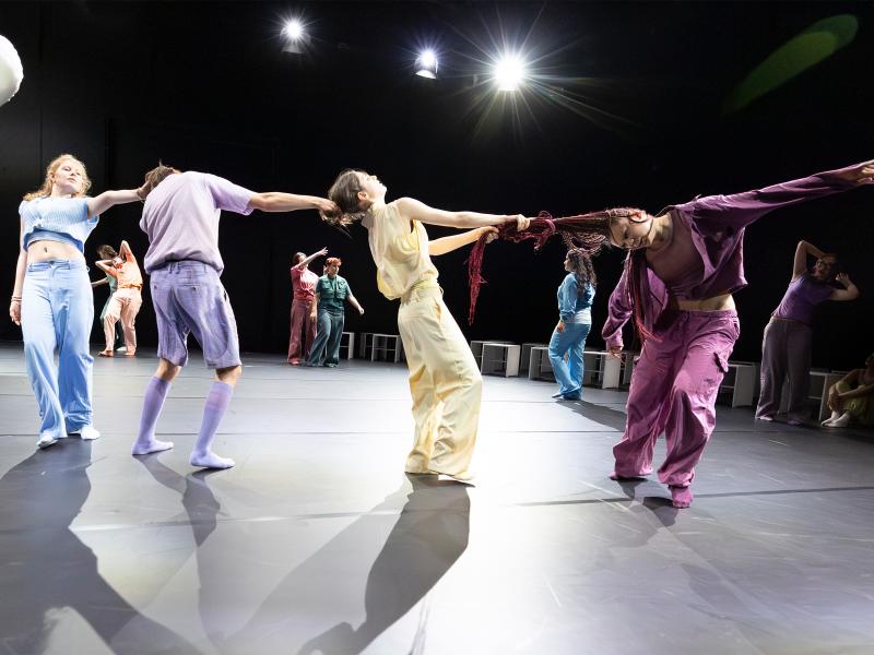 Vier Tänzer*innen stehen in einer Reihe und reißen sich gegenseitig an den Haaren. Mit Haaren und Armen entsteht dabei eine Menschenkette. Im Hintergrund tanzen weitere Tänzer*innen. 