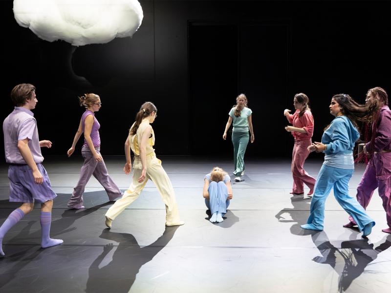 Eine Tänzerin sitzt auf dem Boden und vergräbt das Gesicht in den Knien. Die anderen Tänzer*innen laufen verteilt von allen Seiten auf sie zu. Über ihnen ist eine Wolke.