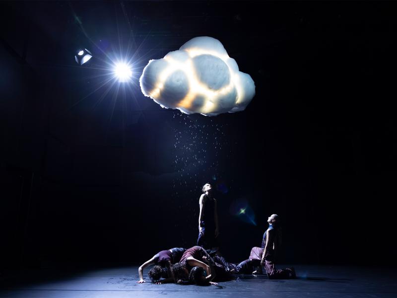 Einige Tänzer*innen stehen und krabbeln unter einer Wolke, die regnet. Einige schauen nach oben in die Wassertropfen.