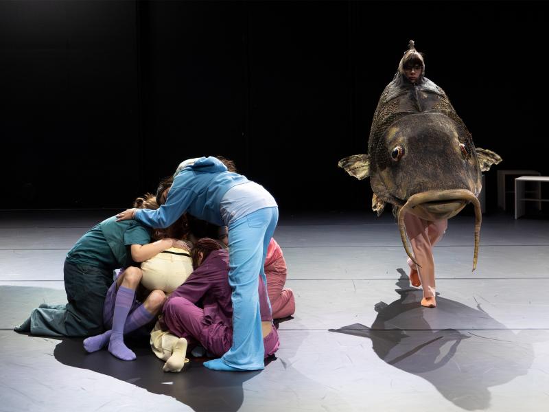 Eine Gruppe von Tänzer*innen bildet einen Menschenhaufen. Daneben läuft ein Tänzer in einem Fischkostüm vorbei. 