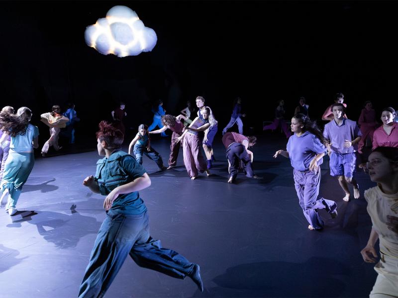 Die Tänzer*innen rennen auf der Bühne im Kreis. Über ihnen eine Wolke, die leicht leuchtet. 