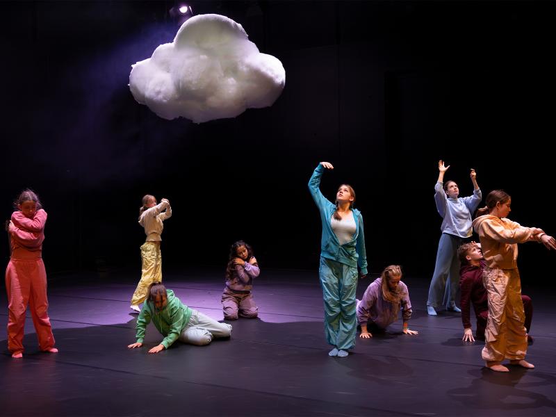 Sechs Tänzer*innen stehen, sitzen und liegen unter einer Wolke, die regnet. Einige schauen nach oben in die Wassertropfen.