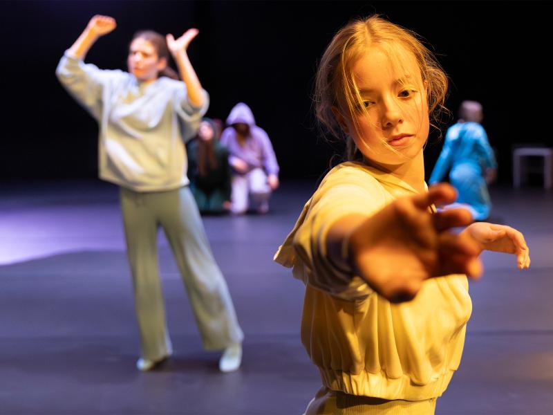 Eine Tänzerin steht im Vordergrund. Sie streckt eine Hand nach vorne und blickt dabei nach unten. Ihre Haare sind in ihrem Gesicht. Im Hintergrund sind weitere Tänzer*innen zu sehen. 