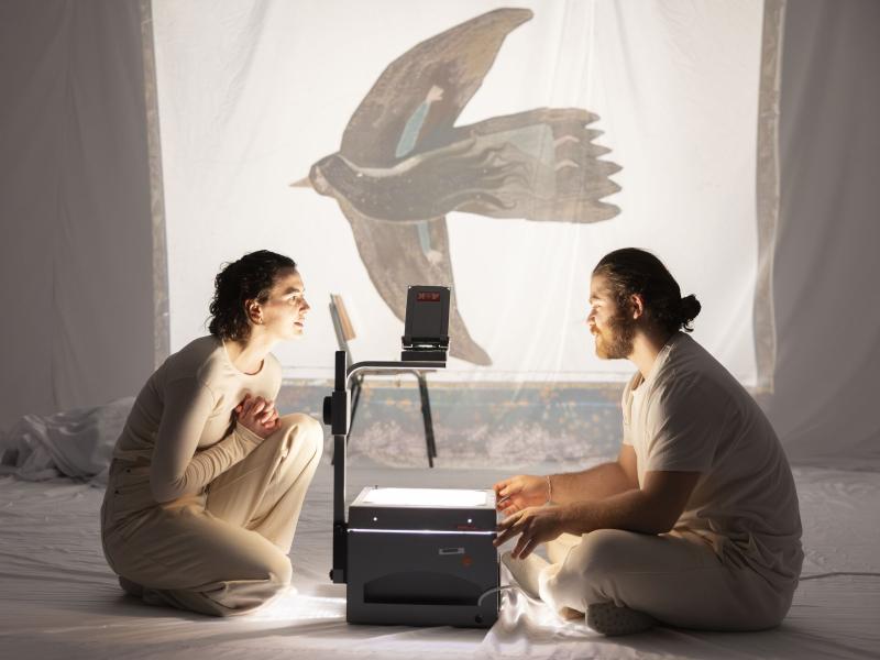 Victoria Kraft und Parsa Yaghoubi Pour sitzen sich gegenüber auf dem Boden. In ihrer Mitte steht ein Overheadprojektor, der auf ein weißes Tuch im Hintergrund ein gemaltes Bild von einem Vogel projiziert. Im Inneren des Vogels ist eine Frau mit langen Haaren gezeichnet.