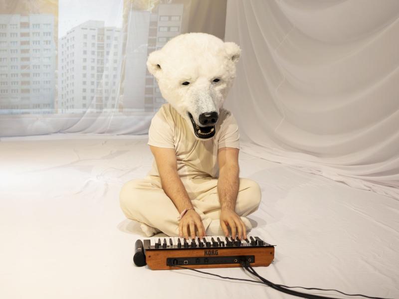 Parsa Yaghoubi Pour sitzt auf dem Boden im Schneidersitz und spielt auf einem kleinen Keyboard. Auf seinem Kopf trägt er eine große Eisbärenmaske. Um ihn herum sind weiße Tücher aufgespannt, die Falten schlagen.