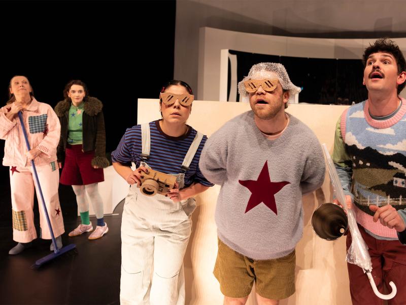 Das Ensemble steht vor großen weißen Bühnenbildteilen. Christine Ochsenhofer und Victoria Kraft stehen im Hintergrund links im Bild. Anastasia Lara Heller, Parsa Yaghoubi Pour und Silvio Kretschmer stehen vorne. Sie schauen erstaunt in die Ferne, Richtung Publikum. Parsa und Anastasia tragen beide eine große Brille aus Pappe. Anastasia hält eine Kamera in der Hand, sie ist ebenfalls aus Pappe. Silvio hält einen durchsichtigen Regenschirm und ein kleines Megafon in der Hand. 