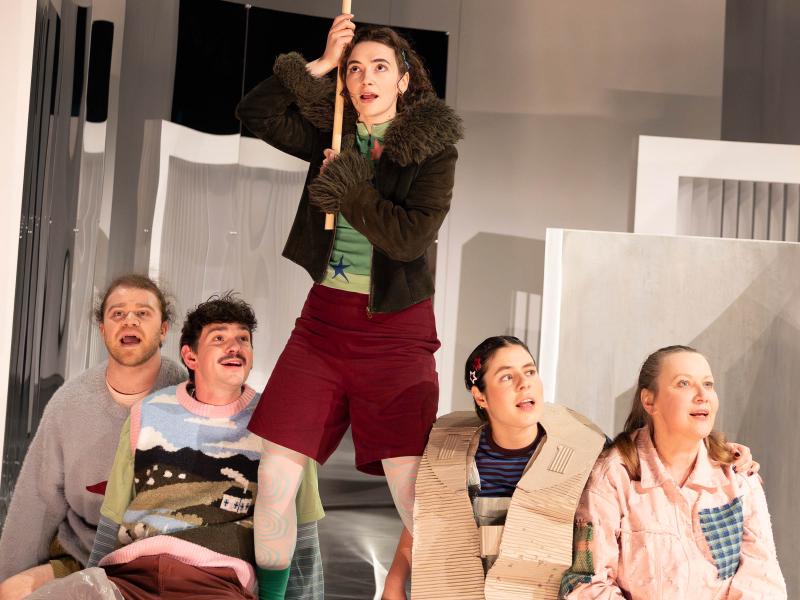 Parsa Yaghoubi Pour, Silvio Kretschmer, Anastasia Lara Heller und Christine Ochsenhofer sitzen vorne an der Bühne. Sie haben den Mund leicht geöffnet und schauen Richtung Publikum. Hinter ihnen große helle Bühnenbildteile, auf ihrem Schoß liegt eine große durchsichtige Plane. Victoria Kraft steht zwischen ihnen und hält eine große Flagge an einem Stab in beiden Händen, die sie in die Höhe streckt. 