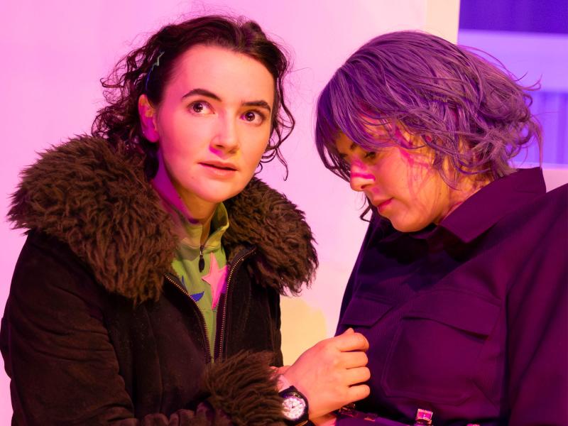 Victoria Kraft und Anastasia Lara Heller stehen sich gegenüber. Während Victoria mit aufgerissenen Augen zur Seite starrt, blickt Anastasia nach unten auf ihre Hände, die an Victorias Armbanduhr fassen. Victoria trägt eine dunkelbraune Felljacke und Anastasia eine lilafarbene Perücke und über eine lilafarbene Anzugjacke einen Gürtel bestehend aus Armbanduhren. 