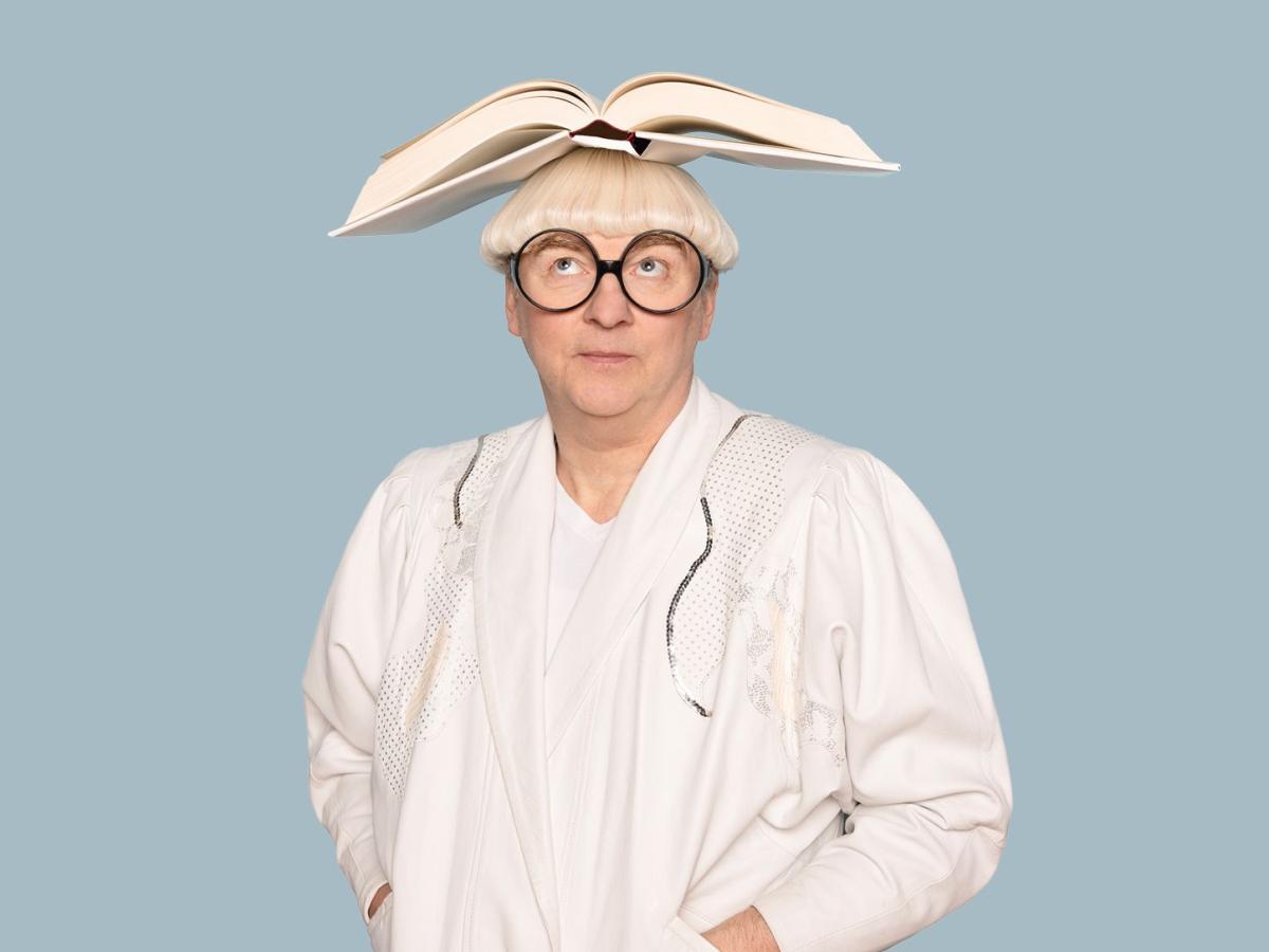 Das Bild zeigt den Schauspieler Hermann Book mit weißer Perücke, großer, runder Hornbrille, einem weißen Tshirt und dadrüber einer weißen Jacke. Auf dem Kopf balanciert er ein offenes Buch.
