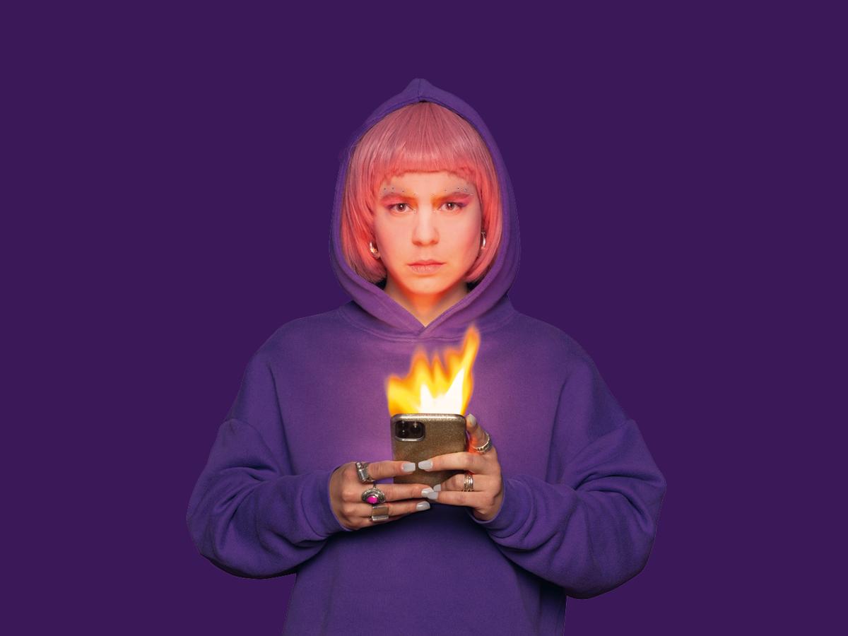 Die Schauspielerin Anastasia Lara Heller steht vor einem violetten Hintergrund. Sie trägt eine pinke Perücke, einen lilafarbenen Hoodie und hat die Kapuze auf. Sie hält ein Handy in beiden Händen, das brennt.