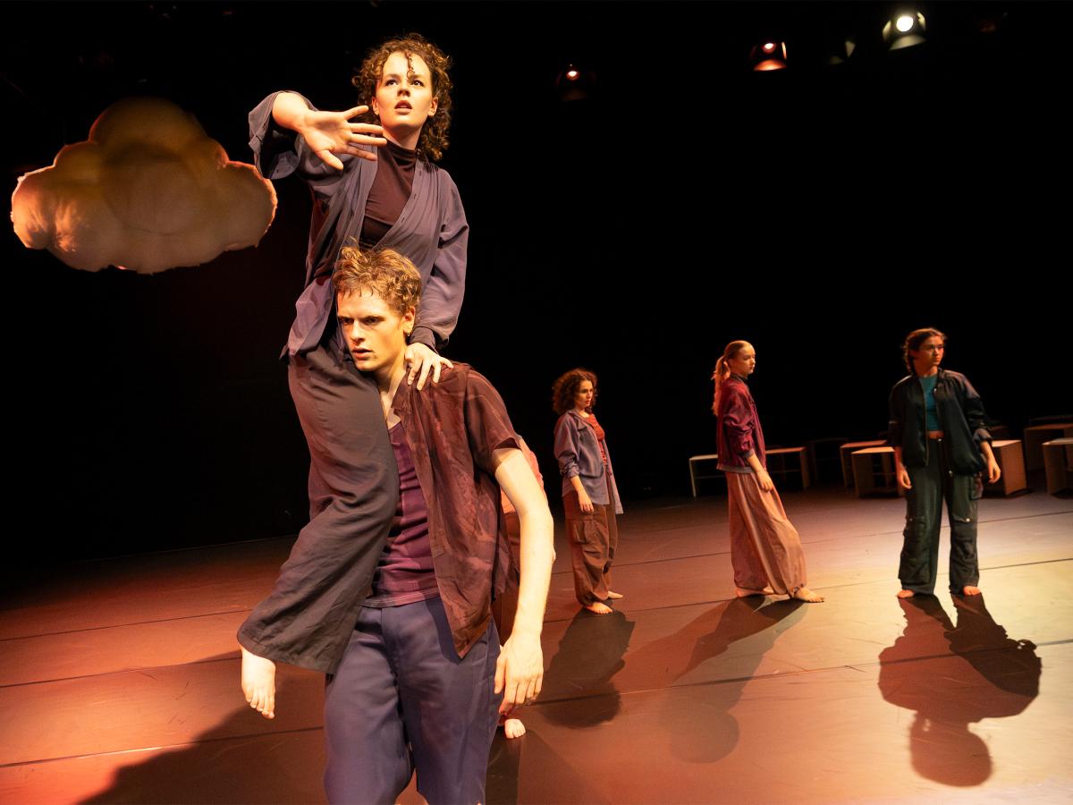 Eine Tänzerin sitzt auf der Schulter eines Tänzers. Sie hält ihre Hand vor sich. Im Hintergrund ist eine Wolke und weitere Tänzerinnen zu sehen. 