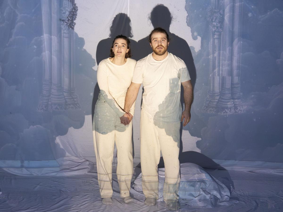 Victoria Kraft und Parsa Yaghoubi Pour stehen nebeneinander und blicken mit offenen Mündern in die Ferne. Victoria hält Parsas Hand mit beiden Händen umklammert. Sie stehen vor einem aufgespannten weißen Tuch, auf das ein gotischer Torbogen projiziert wird, der die beiden umringt. 