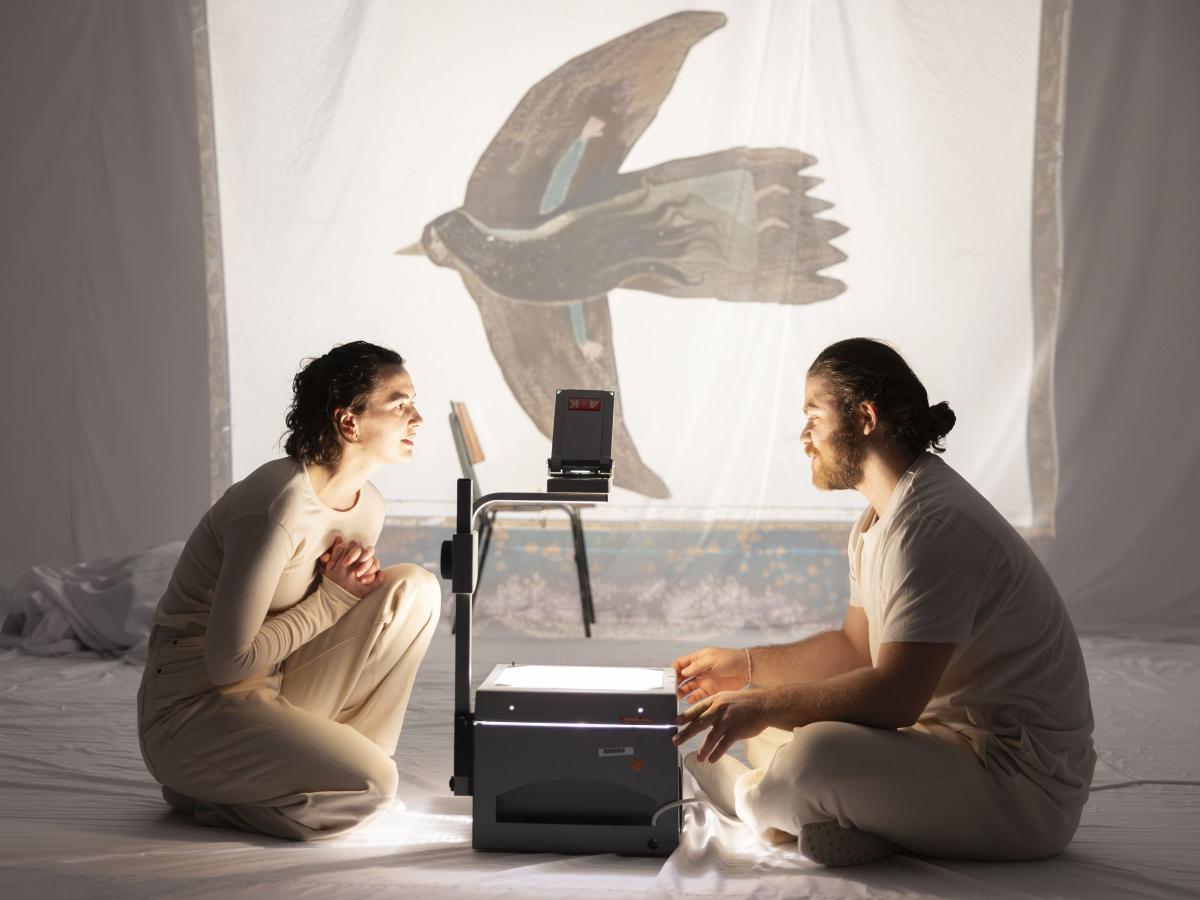 Victoria Kraft und Parsa Yaghoubi Pour sitzen sich gegenüber auf dem Boden. In ihrer Mitte steht ein Overheadprojektor, der auf ein weißes Tuch im Hintergrund ein gemaltes Bild von einem Vogel projiziert. Im Inneren des Vogels ist eine Frau mit langen Haaren gezeichnet.