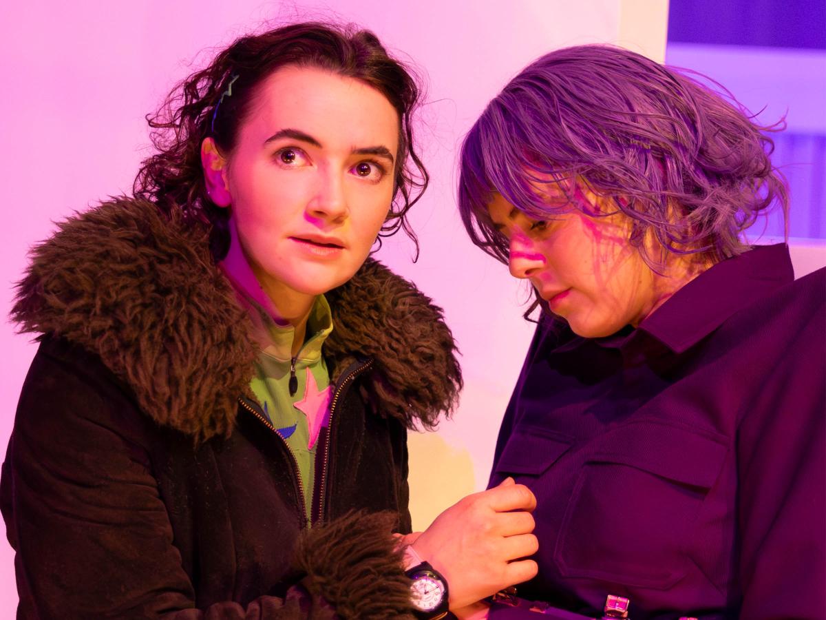 Victoria Kraft und Anastasia Lara Heller stehen sich gegenüber. Während Victoria mit aufgerissenen Augen zur Seite starrt, blickt Anastasia nach unten auf ihre Hände, die an Victorias Armbanduhr fassen. Victoria trägt eine dunkelbraune Felljacke und Anastasia eine lilafarbene Perücke und über eine lilafarbene Anzugjacke einen Gürtel bestehend aus Armbanduhren. 