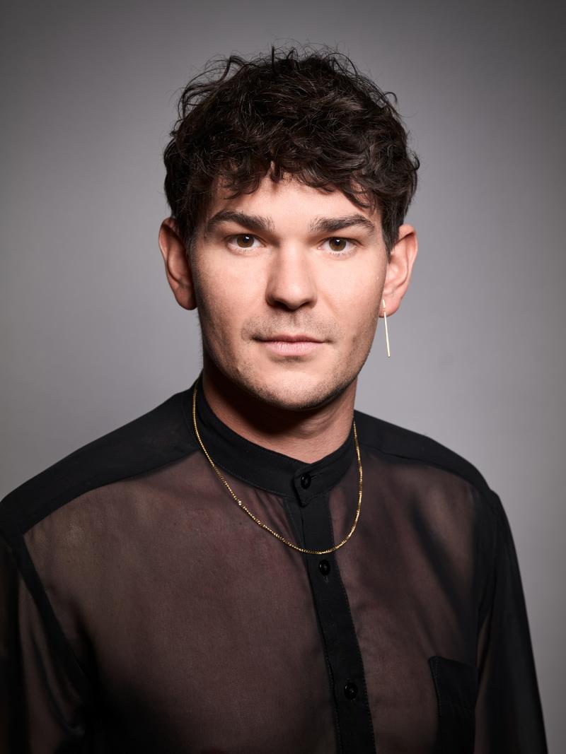 Der Schauspieler Silvio Kretschmer vor einem grauen Hintergrund. Er trägt ein leicht transparentes schwarzes Hemd und eine goldene Kette und schaut neutral in die Kamera.