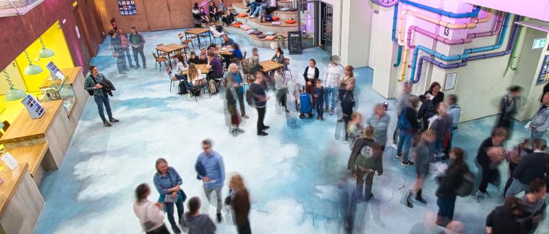 Leichte Vogelperspektive. Das Foyer des Jungen SchauSpielHauses. Die Eingangshalle. Eine bunte Dekoration an der Wand. Die Kasse ist gelb erleuchtet. Menschen stehen im Foyer, sie sind in der Bewegung fotografiert worden und damit unscharf zu sehen.