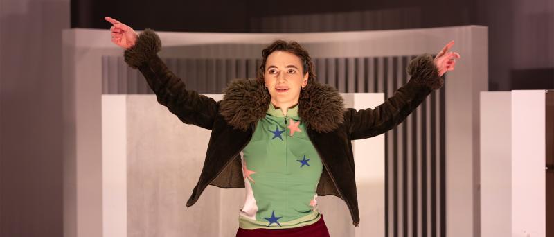 Victoria Kraft steht vor weißen Bühnenbildelementen. Sie trägt dunkle Haare, die zurückgebunden sind. Sie trägt eine Strumpfhose, eine dunkelrote kurze Hose, ein grünes Oberteil, das mit bunten Stern-Applikationen besetzt ist und eine braune Lederjacke mit einem Fellkragen. Sie hat beide Arme rechts und links von sich auf Kopfhöhe ausgebreitet und schaut Richtung Publikum.