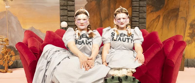Bühnensituation: Christine Ochsenhofer und Anouk Piwek, beide in einem hellen gepunkteten und Kleid mit Puffärmeln sitzen auf einem roten Plüschsofa mit Zacken, die Hände in den Schoß gelegt. Auf ihrer Brust steht „SLEEPING BEAUTY“ in schwarzen Großbuchstaben. Ihre langen blonden Haare sind eingedreht und mit rosafarbenen Bändern festgebunden. Sie tragen Face-Lift-Masken, die Gesicht bis auf Mund und Augen vollständig bedecken. An ihren Füßen befinden sich große grüne Puschen mit schwarzen Krallen daran.