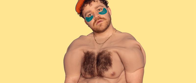 Eine männlich gelesene Person mit gelber Cap und braunen Locken und blauen Augenpads schaut in die Kamera. Über seinem Oberkörper hängt ein menschlicher Oberkörper mit behaarter Brust und Sixpack aus Silikon. Er trägt eine goldene Kette, der Hintergrund ist gelb. 