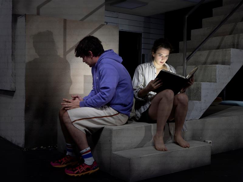 Silvio Kretschmer und Victoria Kraft sitzen auf dem Absatz einer steinernen Treppe. Sie hält ein aufgeklapptes Buch in den Händen, das ihr Gesicht beleuchtet. Silvio sitzt mit dem Rücken zu ihr. 