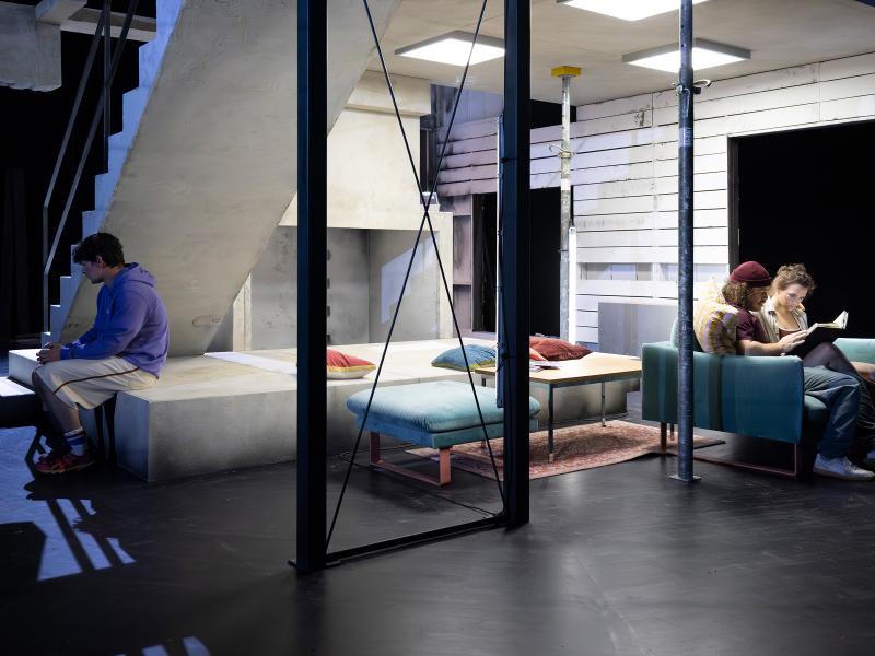 Parsa Yaghoubi Pour und Victoria Kraft sitzen eng beieinander auf einem grünlichen Samtsofa und schauen in ein offenes Buch. Etwas entfernt sitzt Silvio Kretschmer mit dem Rücken zu ihnen. Dazwischen Kissen und eine Sitzbank.