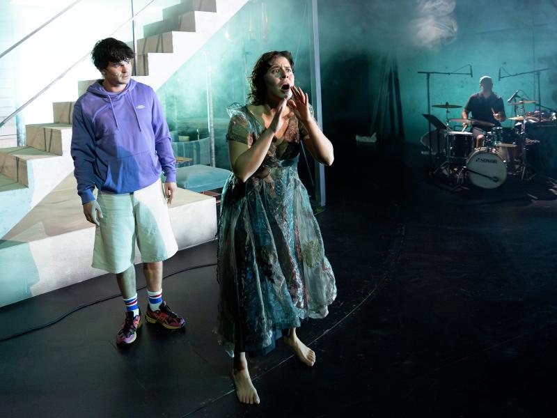 Silvio Kretschmer steht hinter Anastasia Lara Heller und schaut sie an. Sie hat den Mund weit geöffnet und ihre Hände vor dem Gesicht. Ihre Augen sind weit aufgerissen und ihre Augenbrauen zusammengezogen. Im Hintergrund eine Person am Schlagzeug