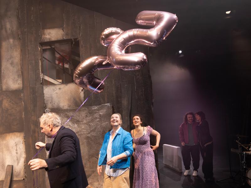 Das Ensemble steht verteilt im Bühnenbild. Hermann Book geht mit Heliumluftballons der Zahlen 2 und 5 vorbei, auf die die anderen lachend aufschauen. Parsa Yaghoubi Pour und Anastasia Lara Heller stehen eng beieinander im Hintergrund.