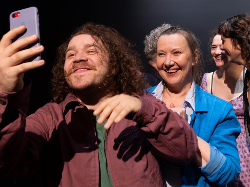 Nahaufnahme vom Ensemble. Parsa Yaghoubi Pour und Christine Ochsenhofer lächeln in ein Smartphone. Die anderen lächeln sich im Hintergrund an und stehen dabei nah beieinander.