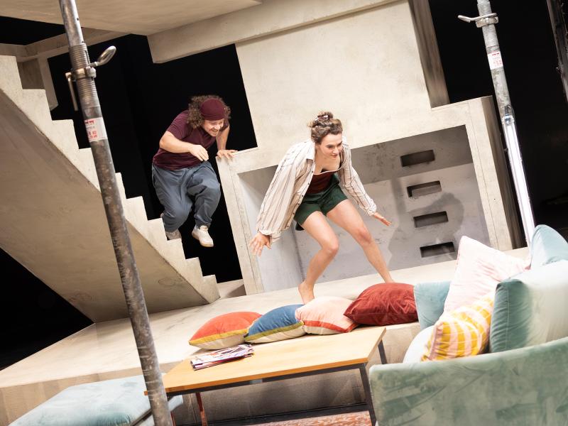 Victoria Kraft und Parsa Yaghoubi Pour bewegen sich lachend durch das Bühnenbild. Im Vordergrund ein grünliches Samtsofa mit Kissen und ein Couchtisch. 