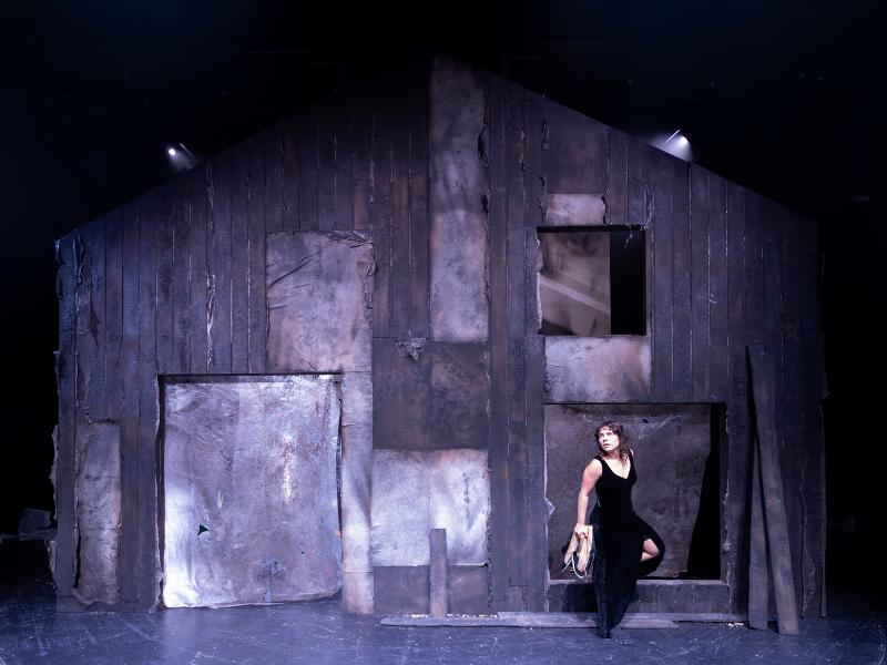 Anastasia Lara Heller steht in einem langen schwarzen Kleid vor einer dunklen, beschädigten Hausfassade. 