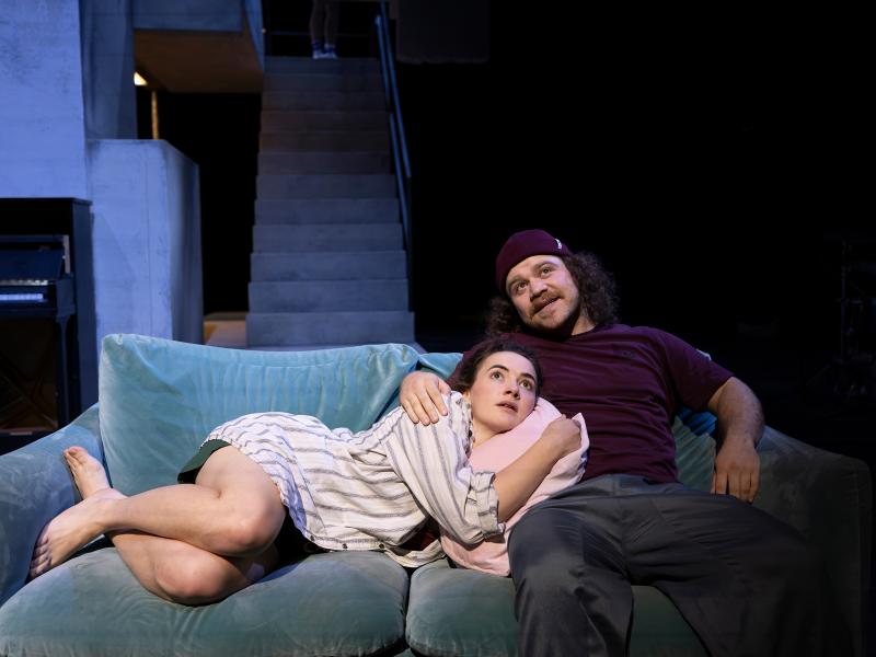 Victoria Kraft liegt auf einem grünlichen Samtsofa in Parsa Yaghoubi Pours Arm. Beide blicken nach oben. Im Hintergrund eine steinerne Treppe. 