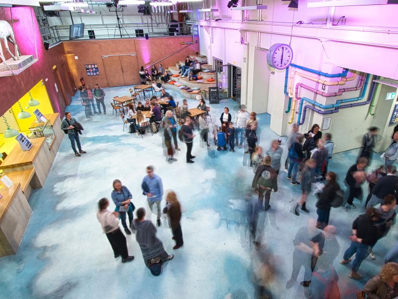Leichte Vogelperspektive. Das Foyer des Jungen SchauSpielHauses. Die Eingangshalle. Eine bunte Dekoration an der Wand. Die Kasse ist gelb erleuchtet. Menschen stehen im Foyer, sie sind in der Bewegung fotografiert worden und damit unscharf zu sehen.