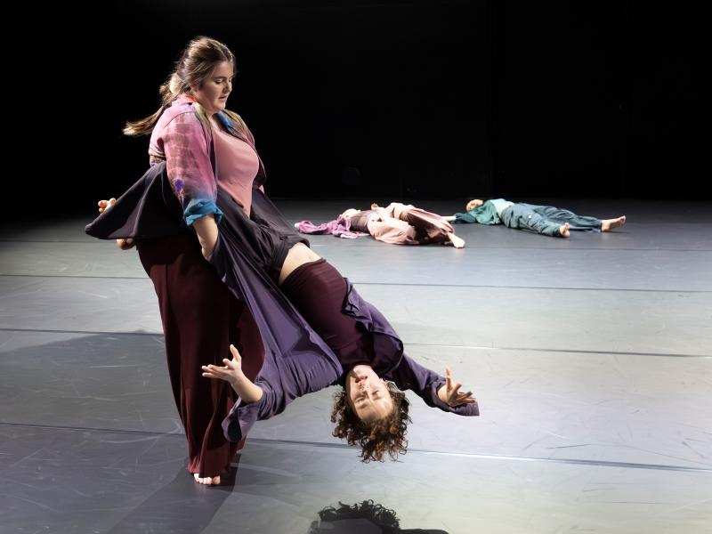 Eine Tänzerin hat ihre Beine um die Hüften einer anderen Tänzerin geschwungen. Die stehende Tänzerin wirbelt die andere herum, die mit dem Kopf nach unten hängt und ihre Arme ausstreckt. Im Hintergrund liegen weitere Tänzer*innen ausgestreckt auf dem Boden.