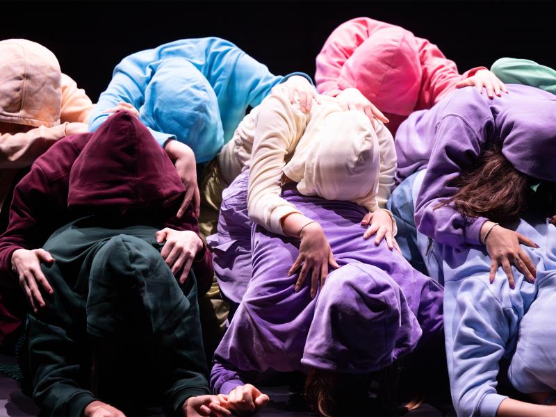 Die Körper der Tänzer*innen bilden eine Mauer, indem sie eingerollt neben- und aufeinander liegen. Man sieht keine Köpfe, alle haben sich Kapuzen aufgezogen. 