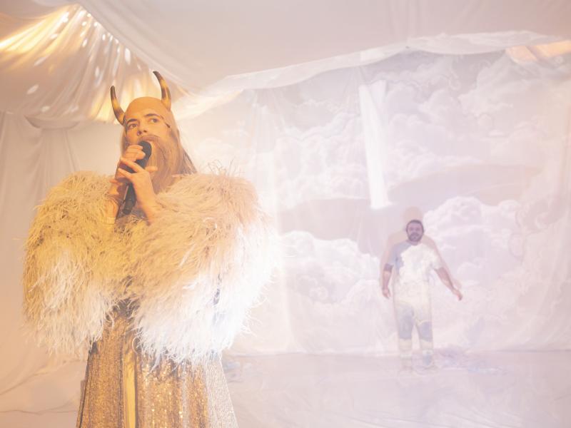 Victoria Kraft steht verkleidet mit einem langen weißen Bart, Felljacke und Hörnern auf dem Kopf im Vordergrund und hält ein Mikrofon in beiden Händen. Parsa Yaghoubi Pour ist im Hintergrund schwach zu erkennen. Beide befinden sich in einem Raum aus weißen Tüchern.