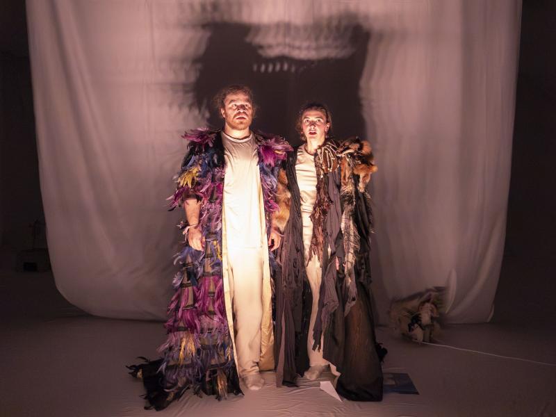 Parsa Yaghoubi Pour und Victoria Kraft stehen dicht nebeneinander und blicken mit offenen Mündern in die Ferne. Sie tragen beide lange Umhänge, Parsas mit bunten Federn und Victorias mit Tierfellen und kleinen Knochen bestückt.