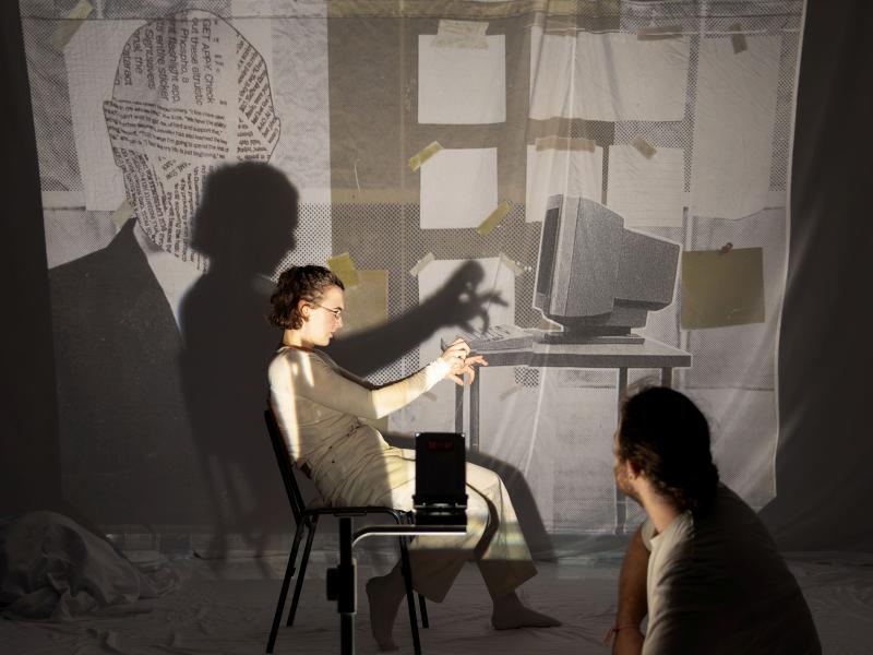 Victoria Kraft sitzt seitlich auf einem Stuhl und macht im Licht eines Overheadprojektors ein Schattenspiel mit ihren Händen. Parsa Yaghoubi Pour schaut ihr dabei zu. Das Bild, dass im Hintergrund auf die Leinwand projiziert wird, zeigt eine Bürowand, an der einige Zettel geklebt sind, und einen Tisch mit einem Computer. 