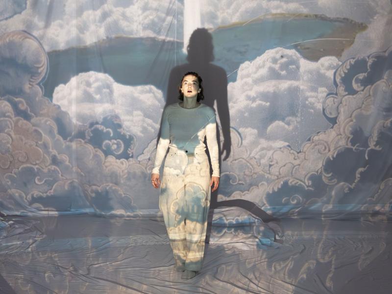 Victoria Kraft steht im Mittelpunkt und starrt mit weit aufgerissenen Augen und offenen Mund nach oben. Sie ist umgeben von weißen Tüchern, auf denen ein Wolkenbild projiziert wird.