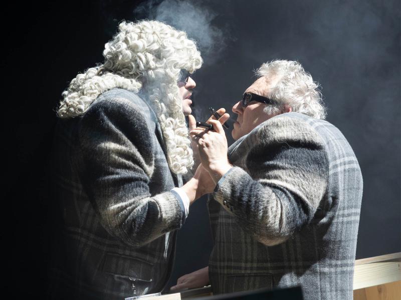 Parsa Yaghoubi Pour und Hermann Book stehen sich gegenüber und schauen sich ins Gesicht. Zwischen ihnen ein Bühnenbildteil, dass ihnen ungefähr bis zur Hüfte reicht. Die Umgebung ist dunkel, nur die beiden Schauspieler stehen im Licht. Parsa trägt eine große, graue Perücke mit Locken. Beide tragen dunkle Sonnenbrillen und den gleichen Anzug, der grau-kariert ist. In ihren Händen halten sie Zigarren-artige Gegenstände, die qualmen. 