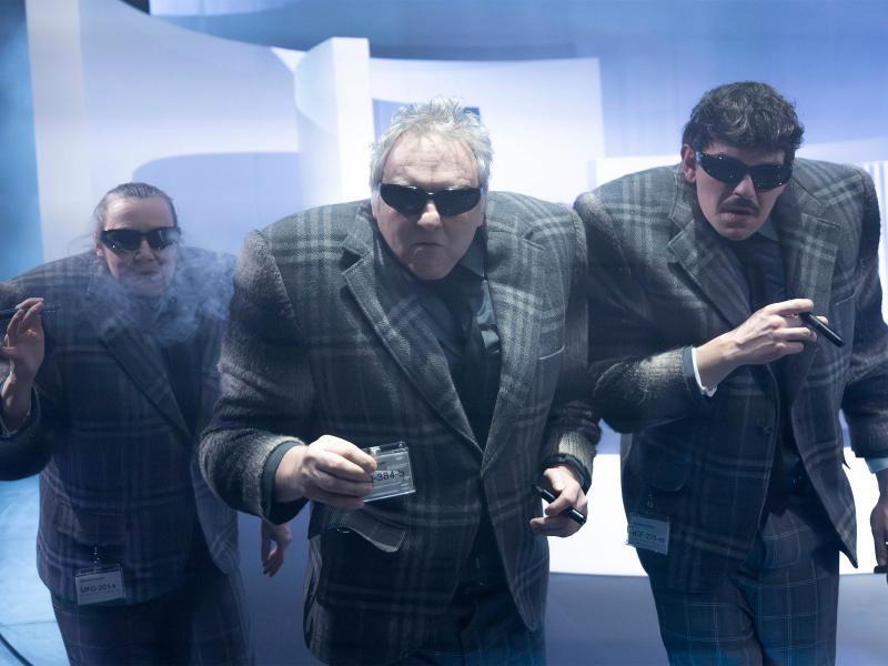 Christine Ochsenhofer, Hermann Book und Silvio Kretschmer stehen nebeneinander in einer Reihe. Unter ihren karierten Anzügen tragen sie sehr breite Schulterpolster. Alle drei haben Sonnenbrillen auf und Zigarren in den Händen. Hermann in der Mitte hält einen Ausweis in der Hand.