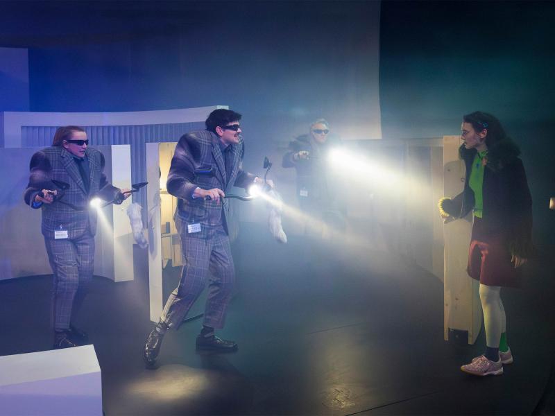 Im Nebel stehen Christine Ochsenhofer, Silvio Kretschmer und Hermann Book in der Mitte der Bühne. Sie tragen alle drei karierte Anzüge und darunter sehr breite Schulterpolster. Sie halten Fahrradlenker mit Lampen in den Händen, die durch den Nebel scheinen und damit Victoria Kraft ins Visier nehmen, die hinter einer Mauer hervorschaut.