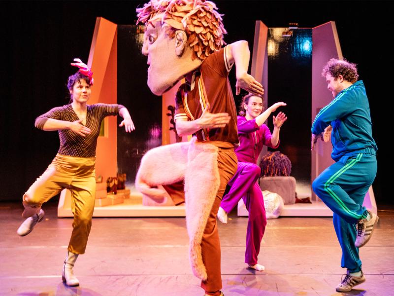 Bühnensituation: Das Ensemble trägt bunte, einfarbige Kleidung in lila, gold, blau und lila. Alle tanzen. Eine Person trägt eine übergroße Kopfmaske mit Gesicht und Haaren. Im Hintergrund kleine, einfarbige hohe Räume in orange und lila.