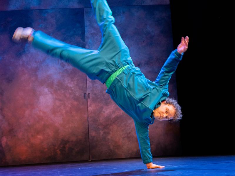 Bühnensituation: Parsa Yaghoubi Pour in einem blauen Trainingsanzug stützt sich mit einer Hand am Boden ab, der restliche Körper befindet sich in der Luft, seine Beine sind ausgestreckt.