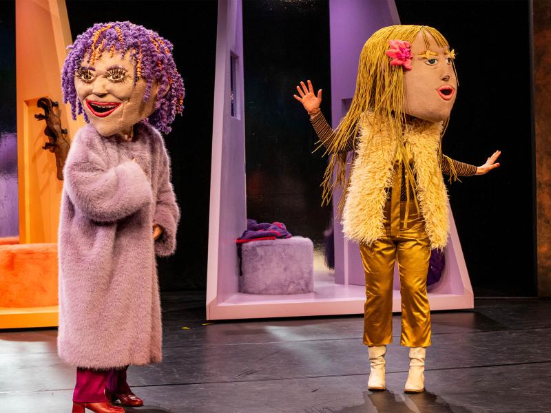 Bühnensituation: Zwei Personen mit aufgesetzten großen Kopfmasken nebeneinander. Die linke Person trägt rote Stiefel, eine rote Hose, einen lilafarbenen flauschigen Mantel, ihre Kopfmaske hat lilafarbene Locken mit orangenen Strähnen und einen geöffneten, lächelnden Mund. Die rechte Person trägt goldene Kleidung zu weißen Stiefeln und eine helle Fellweste, ihre Kopfmaske hat rückenlange blonde Haare mit einer rosa Schleife. Der Mund ist geöffnet. Im Hintergrund angedeutete orange- und lilafarbene Räume.
